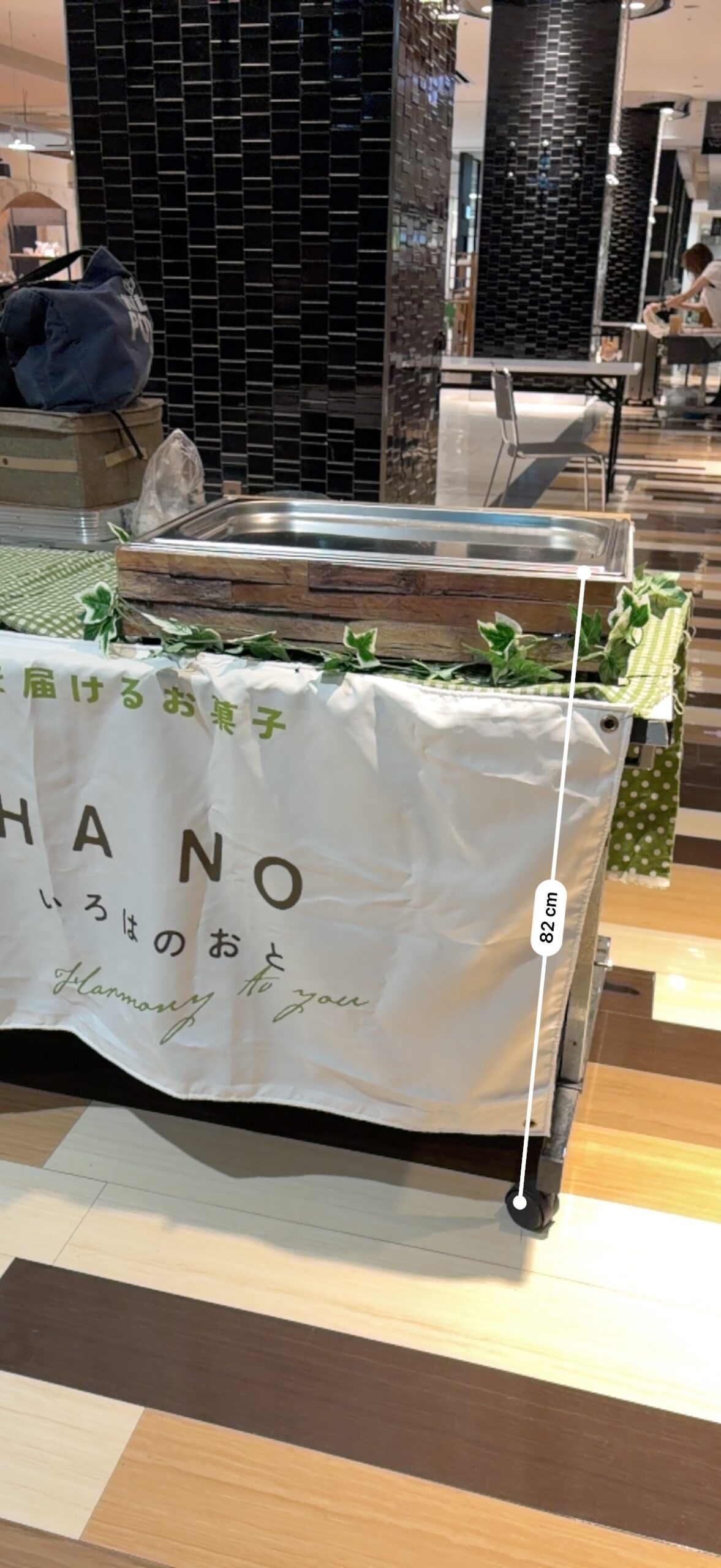 木箱とステンレストレーで商品台を約11cmかさ上げした様子。iPhoneの計測アプリで床から82cmと実測している。
