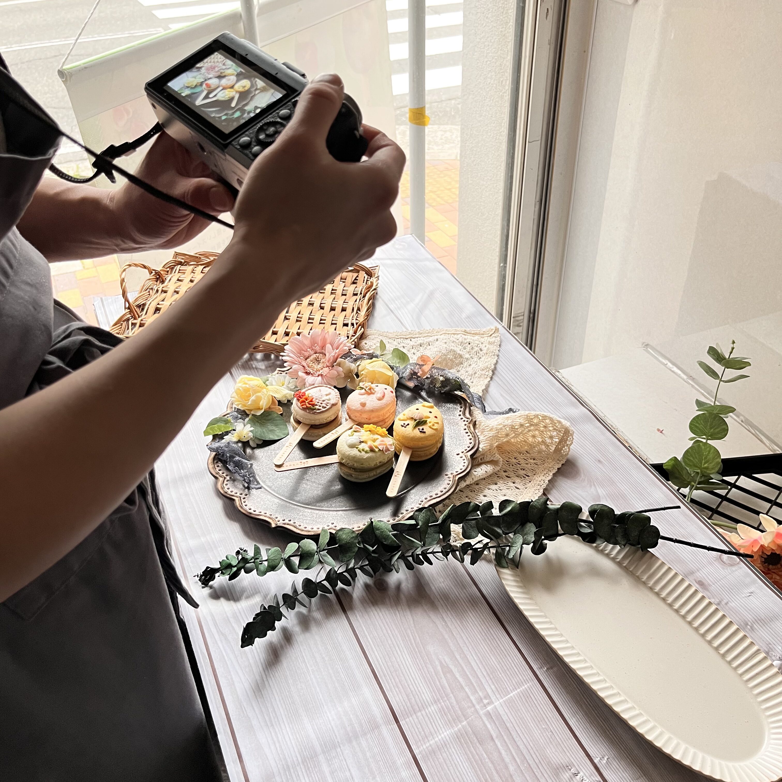 窓ぎわのテーブルで商品写真を撮影している様子。白い木目の背景ボードにレースの布とユーカリを合わせたセッティング。