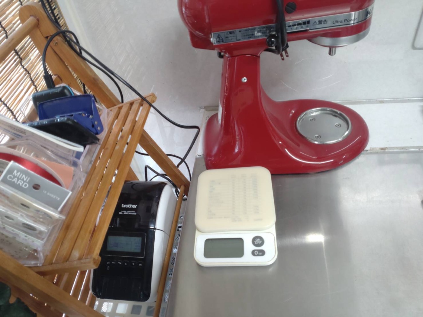 マカロン店の厨房の作業台。木製のラックの下段にブラザーQL-820NWBが置かれ、その隣にステンレスの作業台と赤いキッチンエイドのミキサーが並んでいる。