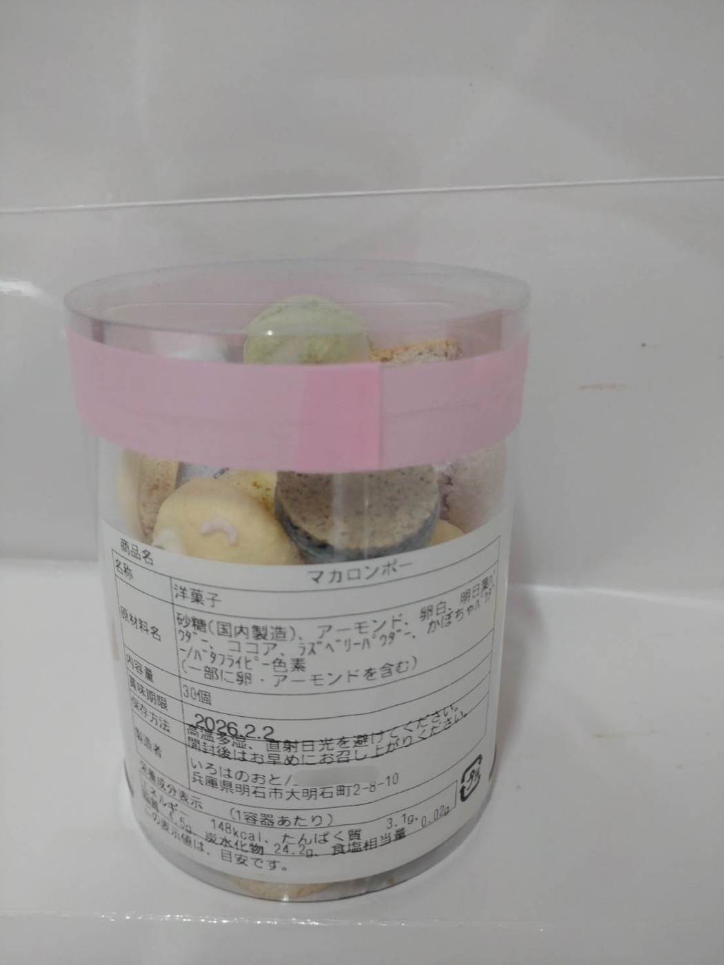 棒付きマカロンの詰め合わせ「マカロンポー」の透明容器に、食品表示シールを貼った状態の写真。ピンクの帯の下に白いラベルが貼られている。