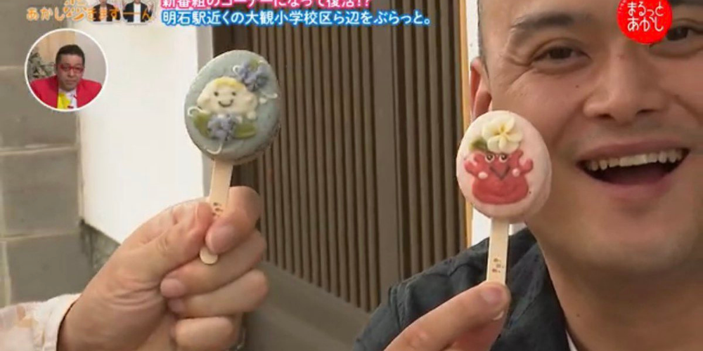 明石ケーブルテレビ「まるっとあかし」の取材で、出演者が棒付きマカロンを2本持っている場面。