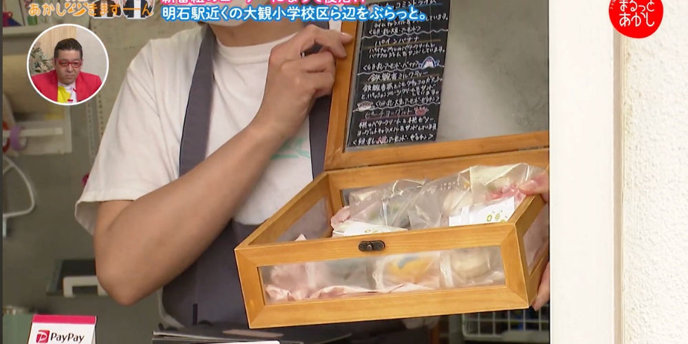 明石ケーブルテレビ「まるっとあかし」の取材で、店頭で焼き菓子の入った木製のケースを見せている場面。