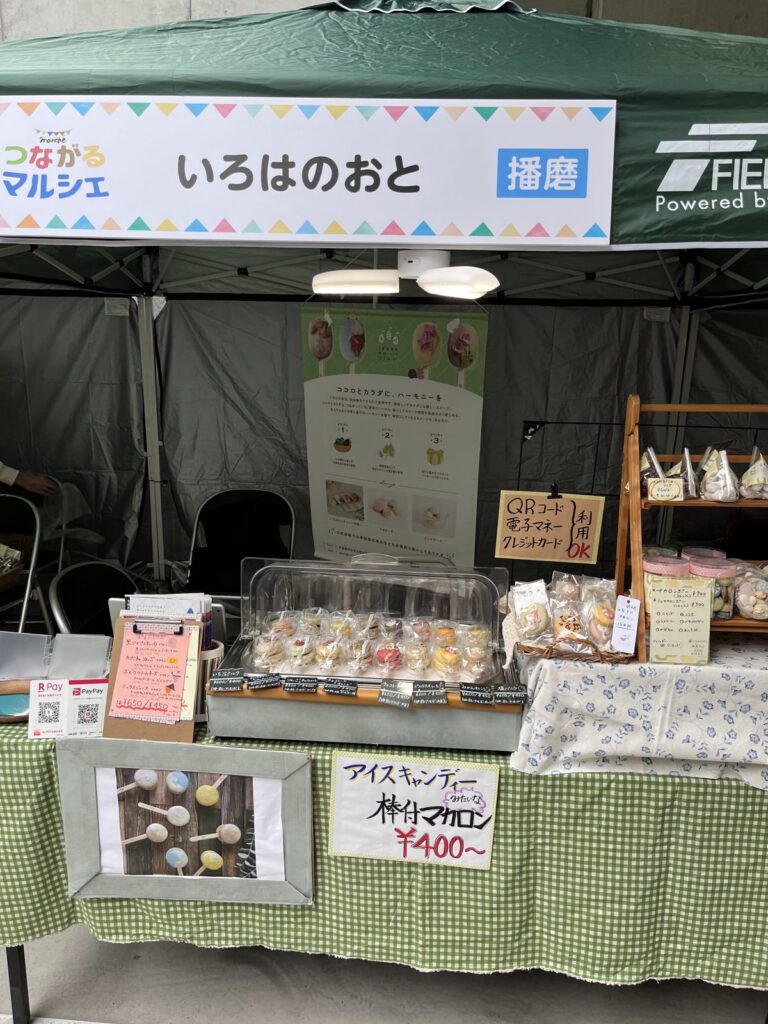 2024年11月の屋外マルシェ。自前のテントに主催者の看板が付き、テーブルには透明のふたの保冷ショーケースで商品を並べている。
